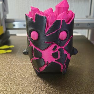 May include: A 3D printed cylindrical container featuring a black exterior with jagged, cracked patterns revealing a bright pink interior. The top edge is filled with multiple pointed, crystalline structures in a vibrant pink colour.