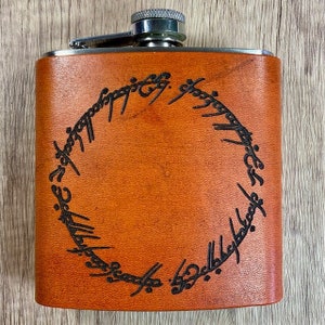 May include: A brown leather flask with a silver top and a black inscription around the center that reads "One Ring to rule them all, One Ring to find them, One Ring to bring them all and in the darkness bind them."