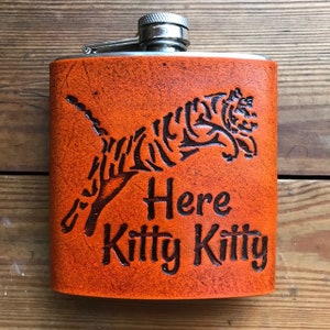 Puede incluir: Frasco de cuero naranja con un tigre grabado en negro y el texto "Here Kitty Kitty".