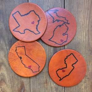 Op de afbeelding: Set van vier leren onderzetters met de contouren van verschillende Amerikaanse staten, elk met een klein rood hartje in het midden. De onderzetters zijn licht oranje.
