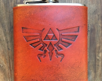 Puede incluir: Una petaca de acero inoxidable con una cubierta de cuero marrón que presenta el símbolo de la Trifuerza de la serie de videojuegos The Legend of Zelda.