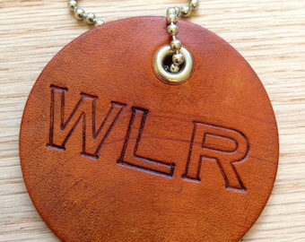Pode incluir: Um chaveiro redondo de couro castanho com uma corrente prateada e um anel de metal. O chaveiro tem as letras "WLR" gravadas.
