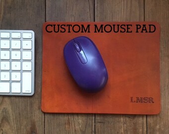 Pode incluir: Um tapete de rato de couro castanho com o texto "CUSTOM MOUSE PAD" em letras pretas e as iniciais "L.M.S.R." em letras brancas. Um rato de computador roxo está no tapete.