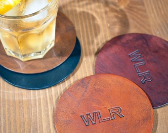 May include: Three round leather coasters with the initials "WLR" embossed on each. The coasters are brown, dark brown, and a reddish brown colour. The coasters are on a wooden surface.