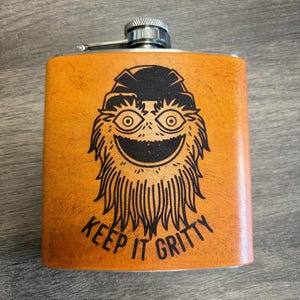 Op de afbeelding: Een rechthoekige, oranje leren heupfles met een roestvrijstalen dop. De fles heeft een zwarte afbeelding van een stripfiguur met de tekst "KEEP IT GRITTY". De fles is ontworpen voor het vervoeren van vloeistoffen.