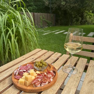 May include: A round wooden serving tray containing cubed yellow cheese, sliced salami, rolled prosciutto, and a small glass bowl of green olives, accompanied by a clear stemmed wine glass filled with white wine on a slatted wooden table.