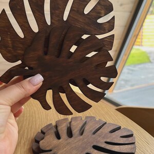 Pode incluir: Dois porta-copos de madeira castanho-escuro em forma de folhas de monstera. Cada porta-copos apresenta um design recortado detalhado que imita as nervuras naturais e as bordas divididas de uma folha tropical, com um acabamento tingido.