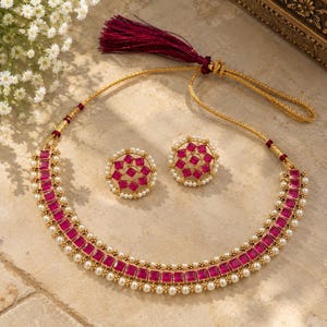 Peut inclure: Un ensemble de collier et boucles d'oreilles assorties en ton doré, orné de pierres magenta de taille carrée et de petites perles blanches. Le collier comprend un long pompon magenta foncé attaché à la chaîne dorée pour un ensemble complet.