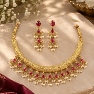 Peut inclure: Un collier doré et une paire de boucles d'oreilles assorties ornés de détails métalliques complexes, de pierres roses en forme de goutte et de petites perles blanches. Le collier présente une bande incurvée texturée avec des pampilles.