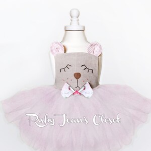 Könnte beinhalten: Ein rosa Tüll-Tutu mit einem braunen Leinen-Oberteil, das ein Bären-Gesicht mit rosa Wangen und einer rosa-weißen Schleife zeigt. Das Oberteil hat rosa Gingham-Ohren. Der Text "Ruby Jean's Closet" ist am unteren Rand des Bildes sichtbar.