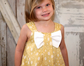 May include: A sleeveless dress in a mustard yellow hue featuring a repeating pattern of white circles. The bodice is accented with a large, solid white bow centered at the neckline, which connects to two wide shoulder straps on each side.