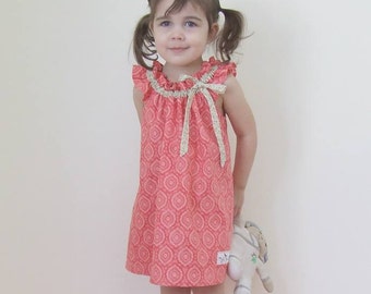 May include: A young girl wearing a coral sleeveless dress with a white and gold pattern. The dress has a ruffled neckline and a bow at the front. She is holding a white stuffed animal.