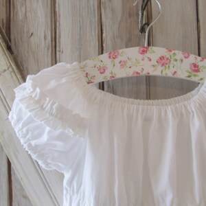 May include: White off-the-shoulder dress with ruffled sleeves hanging on a floral patterned hanger. The dress has a tiered skirt and is made of a lightweight fabric.