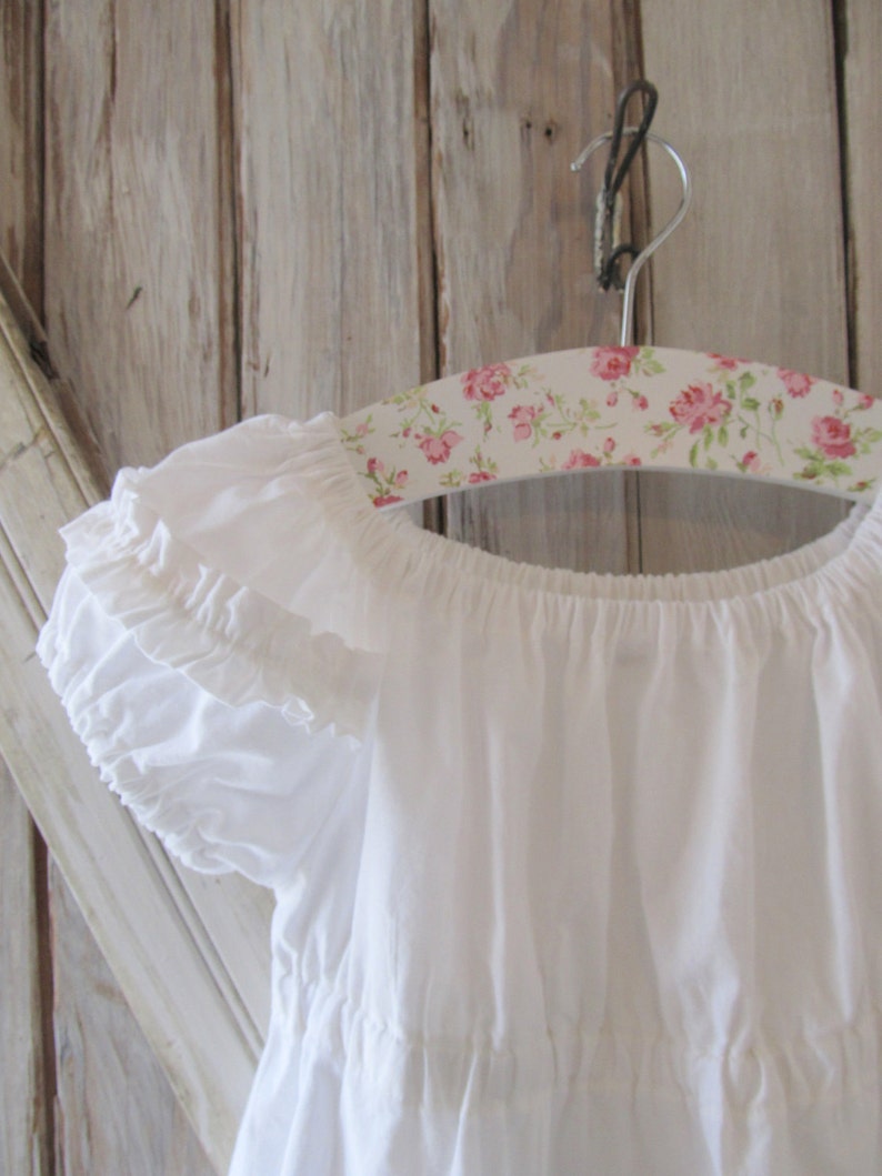May include: White off-the-shoulder dress with ruffled sleeves hanging on a floral patterned hanger. The dress has a tiered skirt and is made of a lightweight fabric.