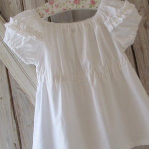 May include: White cotton peasant top with ruffled sleeves and a gathered waist. The top is hanging on a floral patterned hanger.