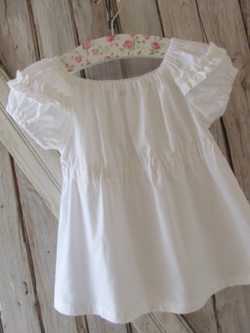 May include: White cotton peasant top with ruffled sleeves and a gathered waist. The top is hanging on a floral patterned hanger.