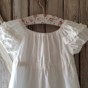 May include: White cotton baby dress with ruffled sleeves and a gathered neckline, hanging on a floral patterned hanger.