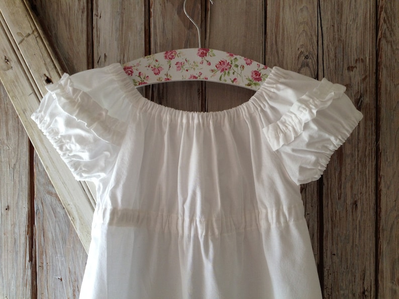 May include: White cotton baby dress with ruffled sleeves and a gathered neckline, hanging on a floral patterned hanger.