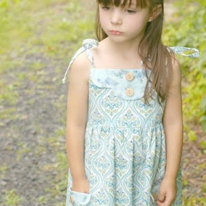 May include: A young girl wearing a light blue and green patterned dress with a white collar and two wooden buttons. The dress has thin straps that tie at the shoulders and two pockets on the sides.