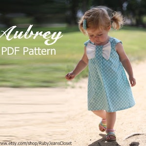 May include: A little girl wearing a light blue and white gingham dress with a large white bow on the front. The dress is sleeveless and has a gathered skirt. The girl is walking on a path with green grass on either side. The text "Aubrey PDF Pattern" is at the top of the image.