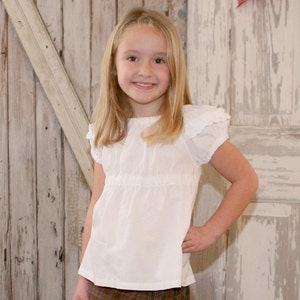 May include: A young girl with long blonde hair is wearing a white, ruffled, short-sleeved blouse with a gathered waist. She is standing in front of a rustic wooden background.