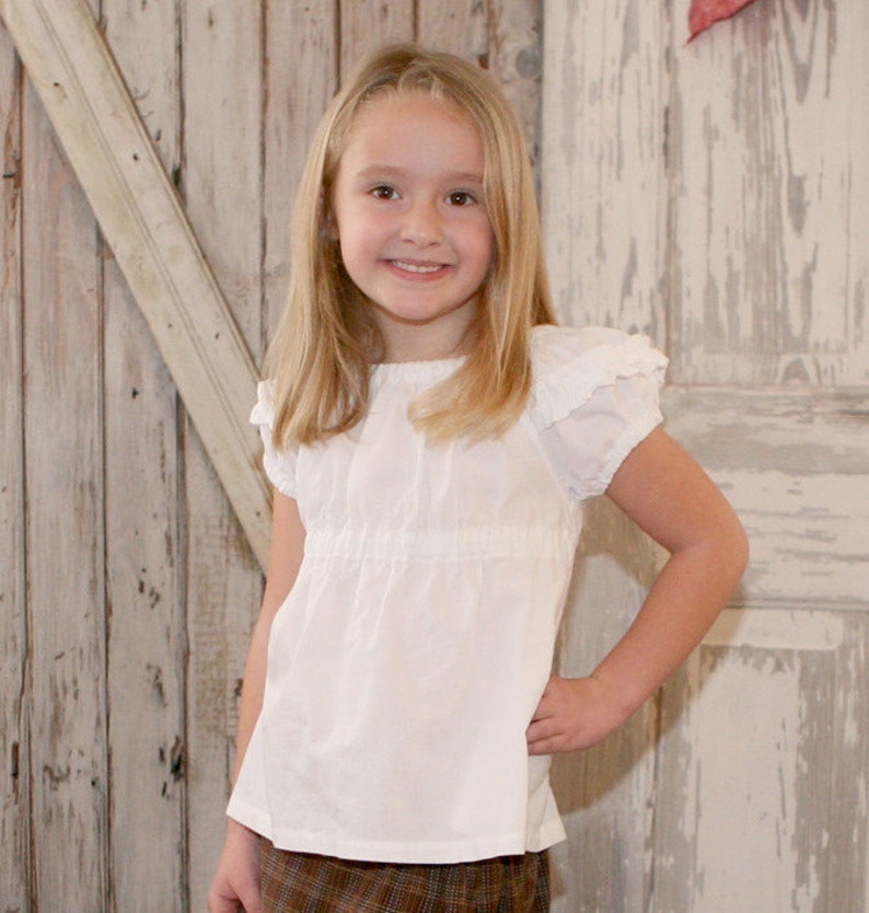May include: A young girl with long blonde hair is wearing a white, ruffled, short-sleeved blouse with a gathered waist. She is standing in front of a rustic wooden background.