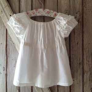 May include: A white cotton blouse with ruffled sleeves and a gathered neckline, hanging on a floral patterned hanger against a wood background.