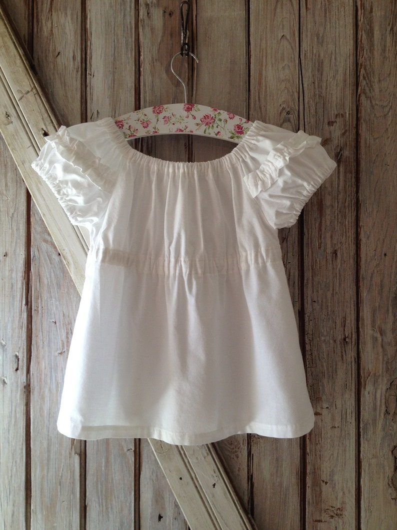 May include: A white cotton blouse with ruffled sleeves and a gathered neckline, hanging on a floral patterned hanger against a wood background.