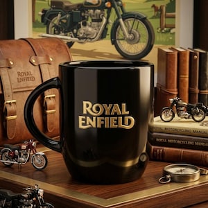 Könnte beinhalten: Ein glänzend schwarzer Keramikbecher mit dem goldenen Royal Enfield-Logo auf der Vorderseite. Der Becher ist umgeben von Miniatur-Motorradmodellen, einer braunen Ledertasche, gestapelten Büchern und einem Kompass auf einer Holzoberfläche.