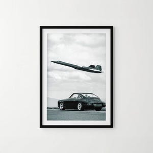 Op de afbeelding: Een zwart-wit ingelijste print met een donkere sportwagen geparkeerd op een verhard oppervlak, met een slank, zwart supersonisch straalvliegtuig dat door een bewolkte lucht erboven vliegt. De compositie toont het voertuig vanuit een achterhoek.
