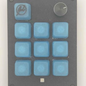 May include: A rectangular black macro pad featuring nine square blue keycaps, one of which displays a circular logo. A single black rotary knob is positioned in the top right corner, and a small power icon is printed at the bottom centre.