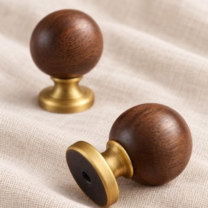 Puede incluir: Dos pomos esféricos para armarios con una cabeza de madera marrón oscuro de grano visible y una base de metal de latón cepillado. La base tiene una placa de montaje circular con un orificio central para tornillo visible en la parte inferior.