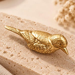 May include: A gold-coloured metal bird figurine with detailed wing and feather textures. The object measures 5.7 cm in length by 1.9 cm in height. The bird is depicted in a resting position with its head turned slightly to the side.