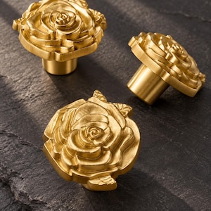 Pode incluir: Três puxadores de armário de metal na cor dourada, cada um com o formato de uma rosa em flor com pétalas em camadas. Os puxadores apresentam um acabamento fosco e uma base cilíndrica para montagem, exibindo um design floral detalhado com um botão.