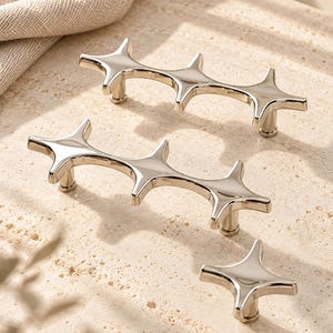 May include: Two elongated silver-toned metal cabinet pulls with a repeating four-pointed star design and one matching single-point star cabinet knob, all featuring a smooth, reflective polished finish and cylindrical mounting posts.