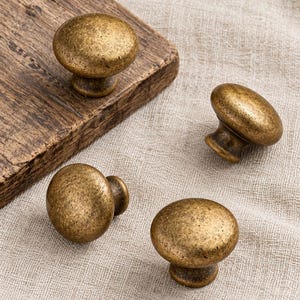 May include: Four round cabinet knobs with a distressed antique brass finish. Each knob features a smooth, domed top and a tapered base, showcasing a textured, weathered metallic surface with dark brown and gold tones throughout the metal.