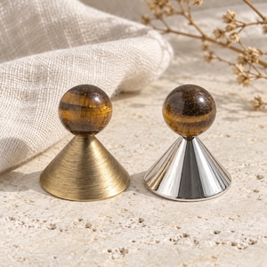 May include: Two decorative objects featuring polished tiger's eye stone spheres atop conical bases. One base has a brushed antique brass finish, while the other has a smooth, reflective silver-tone metallic finish.