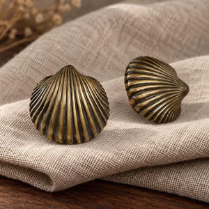 Può includere: Due pomelli per armadietti a forma di conchiglia con una finitura in ottone antico scuro. Ogni pomello presenta una texture dettagliata e scanalata che si irradia dal punto centrale verso il bordo esterno, creando un aspetto realistico.