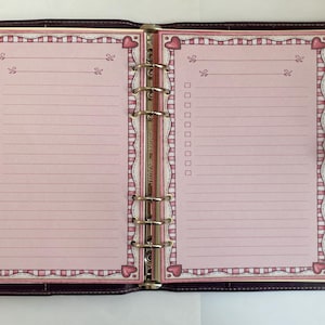 Könnte beinhalten: Ein dunkelvioletter Leder-Ringbuchordner mit sechs Ringen, der auf zwei Seiten hellrosa Papier aufgeschlagen ist. Jede Seite hat einen dekorativen Rand mit rosa Herzen und Streifen, horizontale Linien und eine Spalte mit leeren Kästchen.