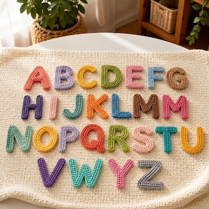Könnte beinhalten: Ein Set aus sechsundzwanzig einzeln gehäkelten Buchstaben des Alphabets, die jeweils in einer anderen einfarbigen Farbe gefertigt sind. Die Buchstaben sind in vier Reihen auf einer strukturierten, cremeweißen Häkeldecke angeordnet und zeigen die Reihenfolge von A bis Z.