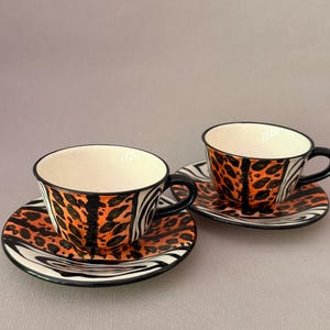Op de afbeelding: Twee keramische theekopjes met bijpassende schoteltjes voorzien van een opvallend patroon van oranje en zwarte luipaardvlekken gecombineerd met zwart-witte zebrastrepen. Elk kopje heeft een zwart handvat en een gladde crèmekleurige binnenkant.