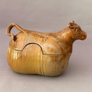 Op de afbeelding: Een beige en bruin geglazuurde keramische pot met deksel in de vorm van een koe. Het object heeft een gebeeldhouwde runderfiguur met een gebogen staarthandvat aan de achterkant, een glad lichaam en een afneembaar deksel in het midden.