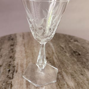 Op de afbeelding: Een helder glazen kelkglas met een geëtst bloemmotief op de kelk. De steel heeft een gefacetteerd ontwerp en de voet is zeshoekig van vorm. Het glas is transparant met witte geëtste details die zichtbaar zijn op de kelk.