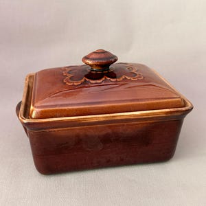 Op de afbeelding: Een rechthoekige botervloot van bruin geglazuurd keramiek met bijpassend deksel. Het deksel heeft een centrale knop als handvat en een decoratieve geschulpte rand aan de bovenzijde. De basis is glad afgewerkt met een donkerdere bruine rand.