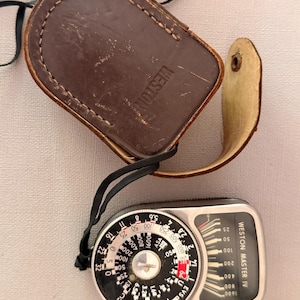 May include: A vintage Weston Master IV light meter with a silver metal casing and a black circular dial featuring white numerical scales. The device is attached to a black strap and is accompanied by a brown leather protective case with Weston embossed.