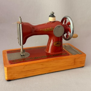 Peut inclure: Une machine à coudre jouet vintage en métal rouge montée sur une base rectangulaire en bois. La machine présente des motifs floraux dorés, une manivelle argentée avec une poignée en bois, une bobine de fil et un logo représentant un cerf.