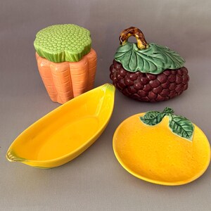 Op de afbeelding: Een set van vier keramische keukenartikelen, waaronder een wortelvormige voorraadpot met een groen deksel, een donkerpaarse druifvormige pot met een groen bladerdeksel, een felgele bananenvormige schaal en een rond geel bord in sinaasappelvorm.