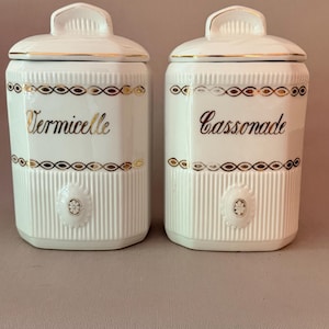 Op de afbeelding: Twee witte keramische voorraadpotten met bijpassende deksels, voorzien van verticaal geribbelde texturen en goudkleurige randen. De ene pot is gelabeld met Vermicelle en de andere met Cassonade in goudkleurig schrift, elk met een ovaal bloemembleem.