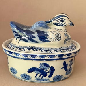 Op de afbeelding: Een ronde keramische pot met deksel, voorzien van een handgeschilderd blauw-wit ontwerp. Het deksel is bekroond met een gebeeldhouwde eendenfiguur. De basis en het deksel zijn versierd met florale motieven, waaronder lotusbloemen en bladeren.