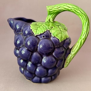 Może przedstawiać: Ceramiczny dzbanek w kształcie kiści ciemnofioletowych winogron. Uchwyt oraz liść na górze są w kolorze jasnej, teksturowanej zieleni. Powierzchnia dzbanka ma błyszczące wykończenie, które podkreśla okrągłe kształty poszczególnych winogron.